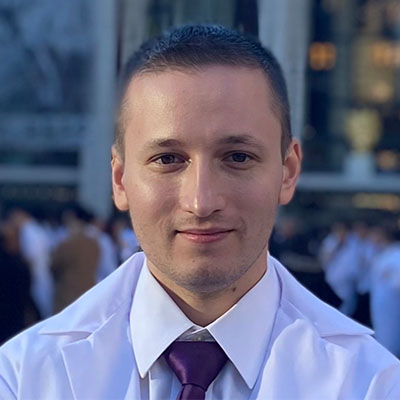 This is a color photograph of a man wearing a white lab coat and a purple tie. He appears to be in an outdoor setting, possibly during the daytime, as suggested by the natural light. The man has short hair, a beard, and is looking directly at the camera with a slight smile.