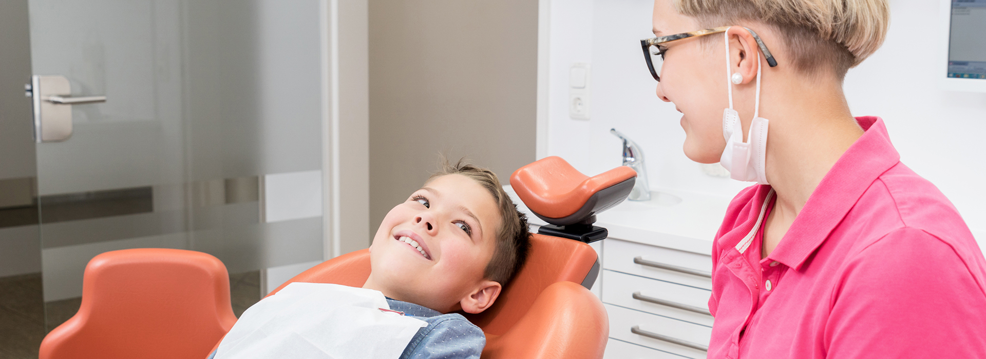 The image shows a dental office setting with a patient in the chair, a dentist standing behind them, and another person seated at a desk.
