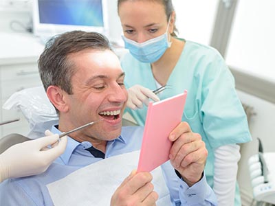The image shows a man in a dental office, smiling and holding up a pink card with the word  Congratulations  written on it. He is seated in a chair with medical equipment around him, wearing a surgical mask, and there s a female dental professional in the background assisting him.