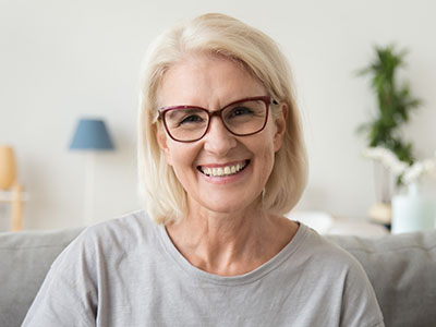 The image shows a woman with short blonde hair, wearing glasses and a gray top, smiling at the camera. She is seated indoors with a neutral-toned background that includes a lamp and some decorative items.