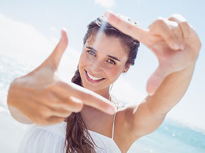 A woman is joyfully taking a selfie with her fingers, smiling brightly against a sunny beach backdrop.