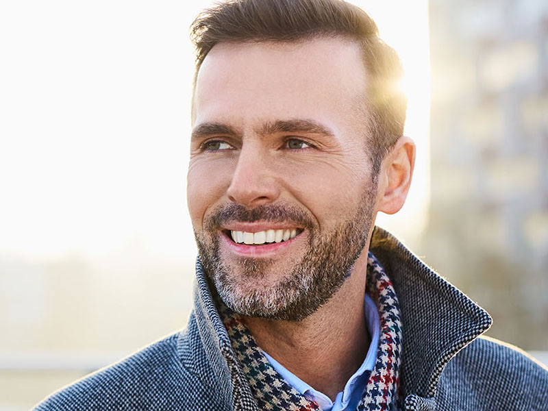 The image shows a man with a beard, wearing a jacket and a checkered shirt, smiling at the camera.